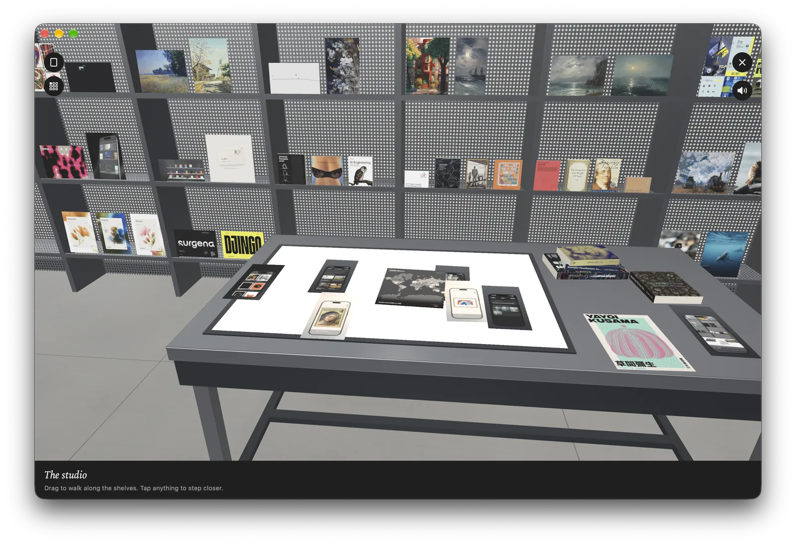 The studio in Atelier on a Mac: grey shelving full of posters and books behind a table with a lit panel holding printed sheets.
